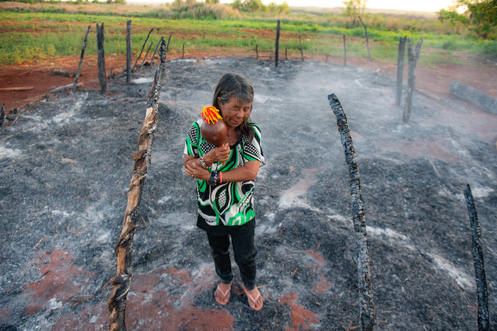 A imagem mostra uma mulher indígena de pé em uma área de terra queimada, abraçando-se em meio a restos de postes de madeira carbonizados e fumaça residual, evidenciando as consequências de um incêndio.